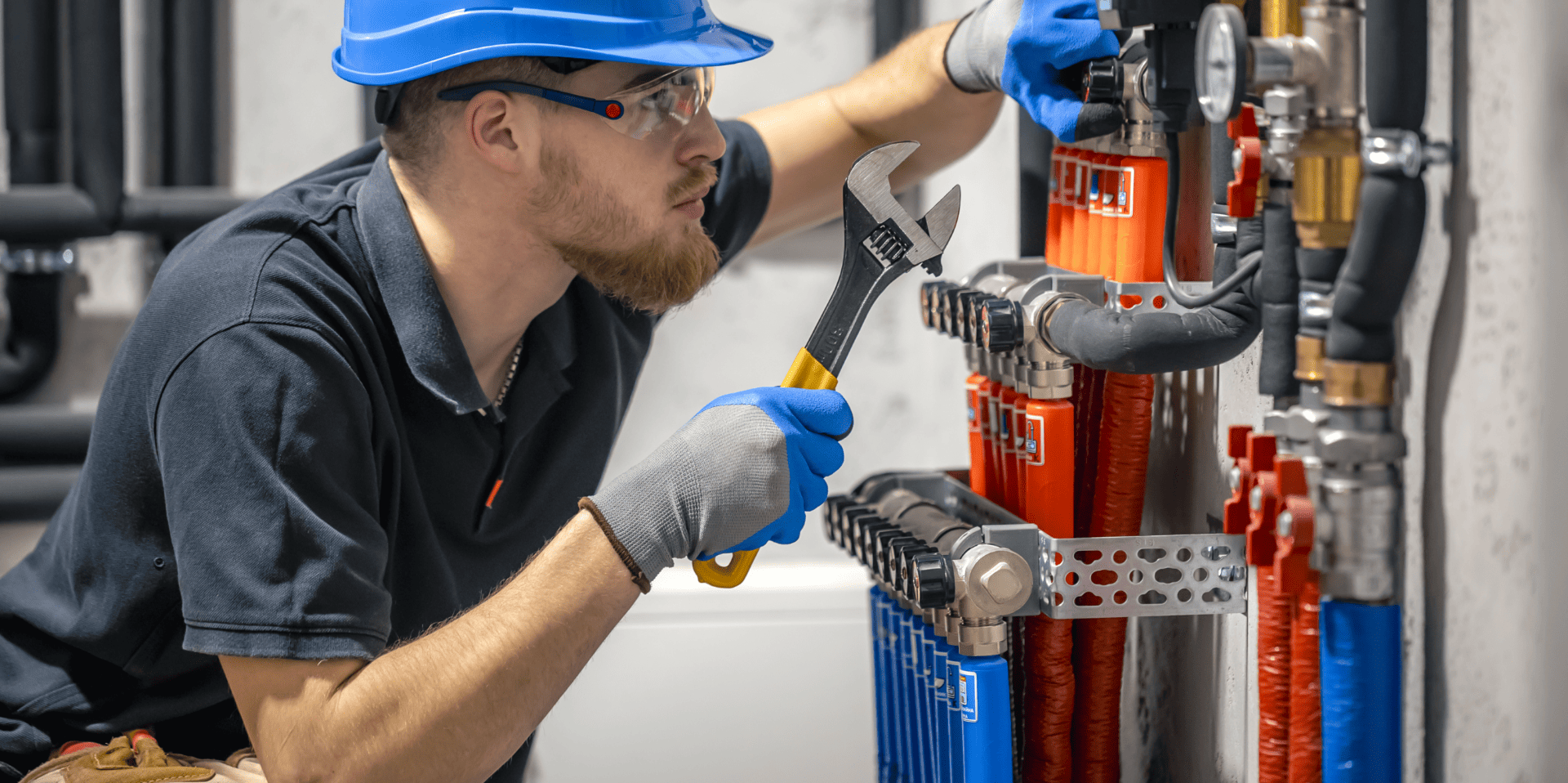 Ein Techniker mit Schutzhelm und Handschuhen arbeitet mit einem Schraubenschlüssel an einer Heizungs- oder Rohrleitungsanlage mit farblich markierten Rohren.